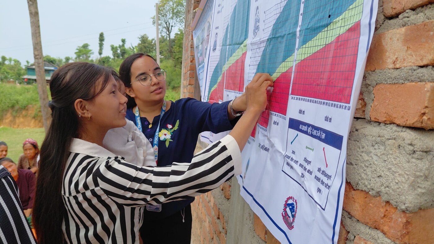 Two community mobilisers marking a child's growth on a wall-mounted growth chart during a courtyard session in Jwalamukhi Rural Municipality, Nepal